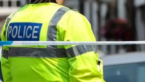 Police officer seen from behind, wearing fluorescent jacket with "police" on the back standing in front of a line of police tape.