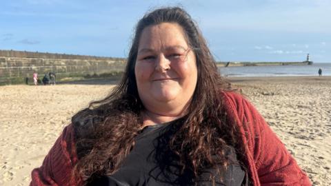 Katie Wotton smiling into the camera on a beach. She's got long, brown hair and is wearing a red cardigan with a black top.