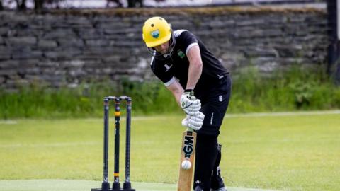 A man wears dark clothes, a yellow helmet and white gloves as he bats a ball in front of three cricket stumps.