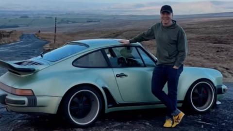 A white man wearing a black baseball hat standing next to an old fashioned Porsche with mountains in the background