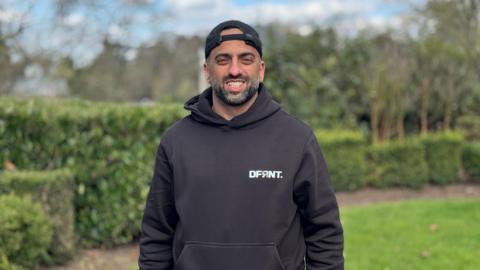A man with a backwards black cap is wearing a black hoody with the letters DFRNT in white on the left side of his chest. He is smiling at the camera and is standing in a park