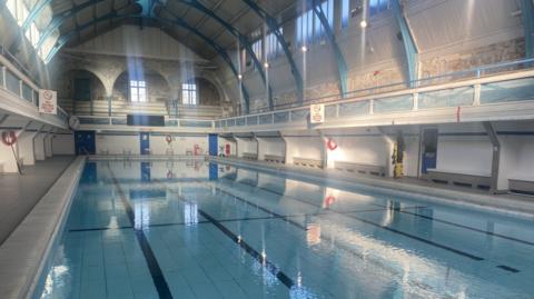 The large refurbished swimming pool, with bright turquoise water and tiled lanes on the bottom. It is inside a Victorian building with white stone walls, arched metal beams and bright white lighting.