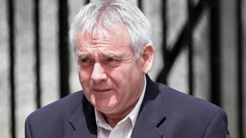 McFarlane with short grey hair looks to the side as he wears a black suit jacket and a white shirt.