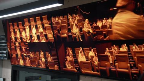 A bank of monitors showing a number of people sitting in an auditorium and looking ahead at a screen. 