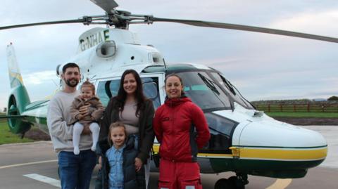 Olivia Brummitt and Dan Atkinson, their daughters and an air ambulance crew member stand in front of Great North Air Ambulance helicopter on a helipad marking.