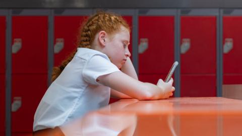 A side view waist-up shot of a female teenager sitting down. high table with her phone. She has her hand on her head looking annoyed and disappointed. She is wearing a full school uniform.