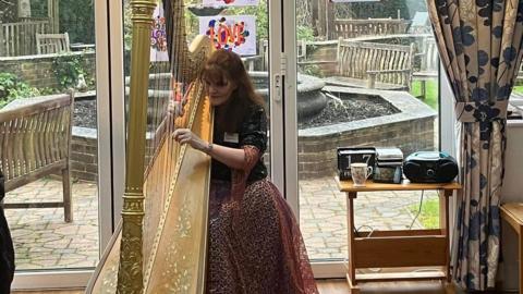 A woman with long red hair dressed in a patterned skirt and black shirt playing a golden harp. Behind her is a water feature and wooden benches.