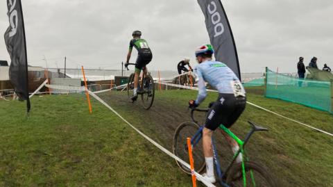Two bike-riders taking part in the 2026 Lloyds National Cyclo-cross Championships are racing up a muddy hill between two tape fences.
