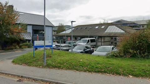Google Street View image of the Mermaid Centre at the Royal Cornwall Hospital. Two buildings - one with light blue cladding and another with grey brick work - are on the site. A car park outside the buildings has several cars parked in it.
