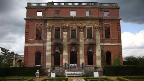 A red brick Georgian building with burn marks on its facade. Firefighters are standing at the bottom of stairs in front of it.