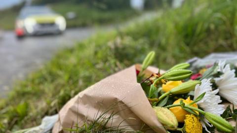 Close-up shot of a bouquet of flowers left at the scene of a helicopter crash in Devon. A police car is blurred in the background.
