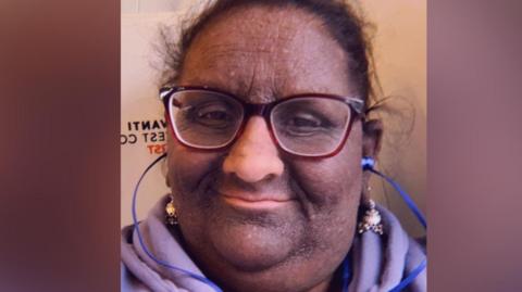 Usha Singh Das has her hair tied back and she is wearing glasses, which are on blue string. She has a lilac coloured scarf on. 