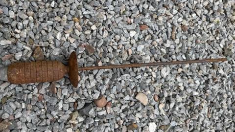 A rusted, cylindrical metal object with ridges and a long thin rod attached lies on a bed of small grey stones. The object appears weathered and corroded. The toe of a dark shoe is visible in the top-left corner, providing a sense of scale.
