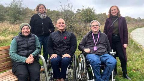 Three men and two women smile at the camera at a nature reserve. Two of the men are in wheelchairs, with the other man sitting on a bench. Both women are standing. They are wearing items including coats, hats and scarves in the autumn weather.