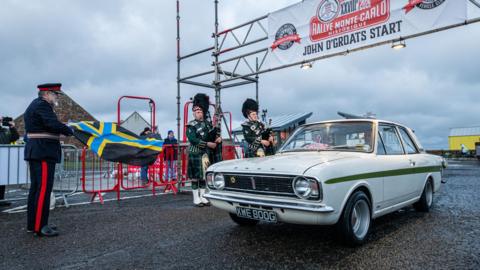 A man wearing a uniform waves the Caithness flag to signal a classic Ford car's departure from John O' Groats. Two pipers stand playing a tune at the start line.