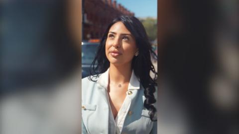 A woman with long black hair, in a light blue suit, looks up while walking in the street