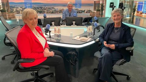  Heather Humphreys and Catherine Connolly in the foreground beside a table with microphones, while presenter David McCullagh is in between them in the background behind three computer screens. Heather Humphreys has short blonde hair and is wearing a red jacket, white blouse and dark trousers. Catherine Connolly has short grey hair and is wearing a dark jacket and trousers and black blouse. David McCullagh has grey hair and is wearing a checked blue shirt.