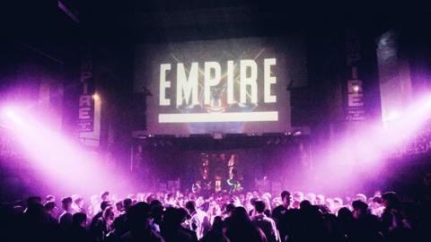 Inside of a nightclub. The dance floor is packed full with people, and purple strobe lights shine down on them from the right and left. Above the dance floor is a screen that says 'EMPIRE' in white.
