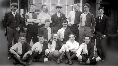 A black and white photograph of Royston Town men's team from 1895. Fourteen players pose together, seven standing, two kneeling and five sitting on the ground. Some are in two-tone football uniform and some in more formal dress. A couple of have mustaches. 