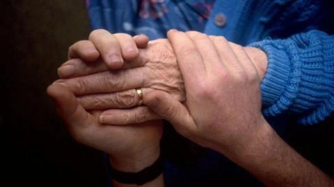 A younger person's hands are clasped around an elderly person's. The elderly person wears a blue jumper.