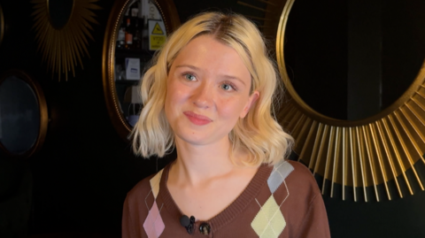A young women with shoulder length blonde hair is smiling. She is wearing a brown cardigan and standing against a wall with two gold mirrors on it