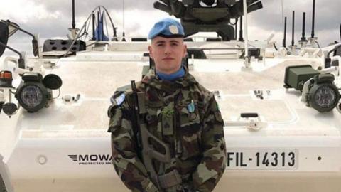 Irish soldier Seán Rooney, wearing camouflage clothes and a blue beret. He is holding a gun and standing in front of an armoured vehicle.