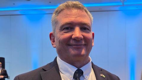 Derek Roberts is wearing a dark suit and a white shirt.