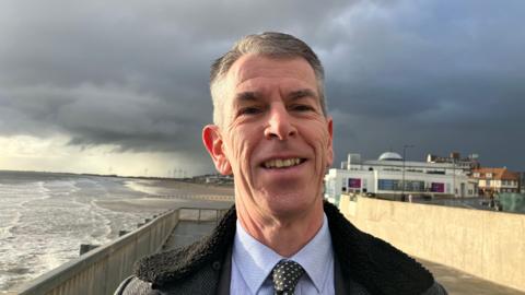 A man with short grey hair is smiling and wearing a black coat, blue shirt and black and white spotty tie. He is standing on a promenade next to a beach. The sky is cloudy.