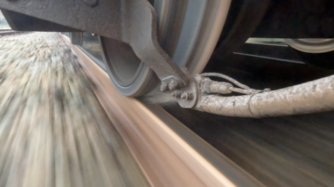 A close up of a train wheel in motion, with a pipe in the foreground spraying sand onto the rail in front of the moving wheel.