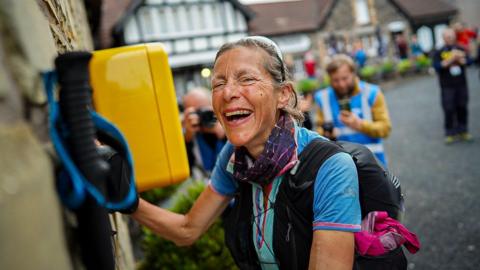 Anna Troup, who is kitted out in trail running attire, including shorts, running rucksack, poles, sunglasses and a sports watch, smiles as she reaches a rest point during the Summer Spine Race. She smiles as she stops in front of race marshals. 