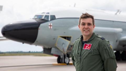 Sqd Ldr Dan Wilkes wearing his flying gear and standing beside an aircraft on a runway with field in the distance. He has his hands behind his back.