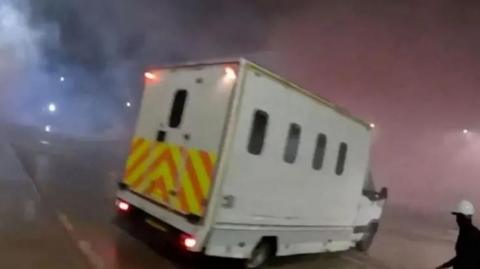 A screenshot of a police van seen on the Elbit Systems UK site. The van is a large white vehicle with small windows and a chevron pattern across the back. A figure wearing black with a white helmet can be seen in the lower right corner of the image holding a long unidentified object.