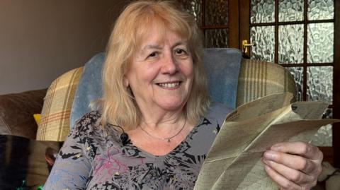 A woman with blonde hair to her shoulders sits in a brown and red tartan chair with her head on a blue cushion. She has a grey top with a black, red and blue flower design outlined on it. In her left hand, which is visible, she holds several pieces of paper with blue writing on them.