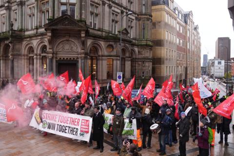 A group of around 100 people with red flags and big white signs are standing on a group. There is also a large amount of smoke in the air. The signs say Unite and Strike Together Win Together. 
