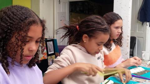 There are three girls sitting in a row at a table making sushi from scratch during a cooking class at Felix Road Adventure Playground in Easton in Bristol.