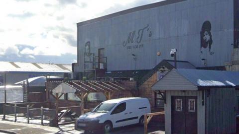 A building with a flat roof and metal cladding along the side. It has a sign that says MJs. In front there is a white van parked next to a seating area with benches and a fence