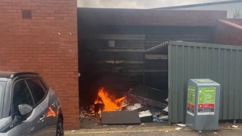 A fire in a bin store, with a metal structure to the right and a metal box. A car is to the left.