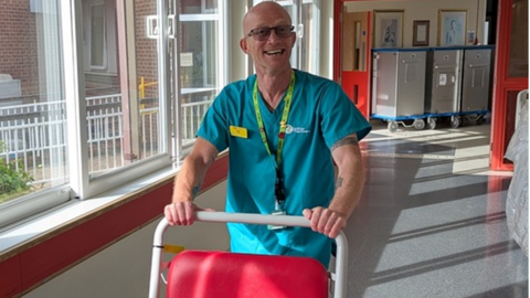 A man in a blue hospital uniform pushes a red trolley with a white frame along a corridor. He wears a name badge and has a lanyard around his neck.