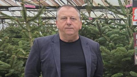 Plants Galore owner Tony Joyner stands in front of dozens of Christmas trees at a shop in Newton Abbot. He is wearing a dark blue blazer and black t-shirt. He has a stern look on his face.
