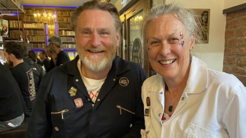 A man and a woman smiling at the camera. He is to the left and has brown hair and a grey beard, she has grey hair tied back. They are both wearing boiler suits - him navy, her white. She has red blue and silver glitter on her face, to the outer side of her eyes. 