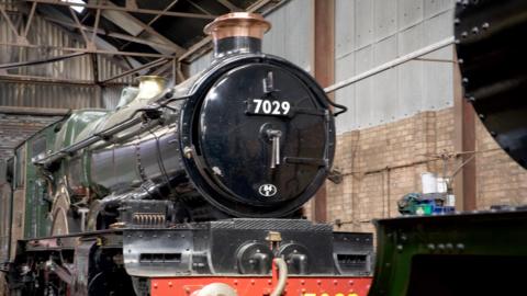 A green steam locomotive in a railway shed. On the front is the number 7029 on a black boiler cover