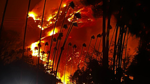 A large mountain can be seen in the distance covered in fires during night time. Large red sections can be seen all the way up the mountain with large plumes of smoke. A road with vehicles travelling along it can be seen at the bottom of the picture.