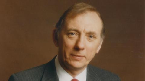 Ken Weetch is looking at the camera. He has short, light brown hair and is wearing a suit jacket, white shirt and a red tie.