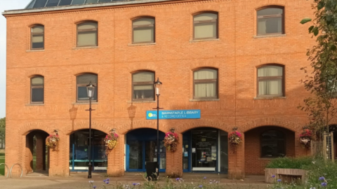 A large, red brick building with three floors. It has five archways on the ground floor. There are five windows on each of the two higher floors. In front of the building are two lampposts