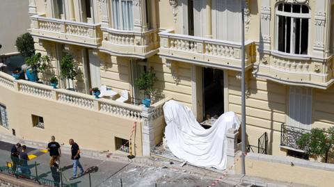 Frontage of a large cream coloured residential building in Monaco with damage to windows and a white sheet accross the front steps and police standing near the front of the building