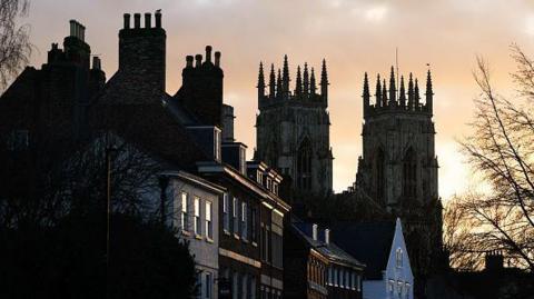 York's historic city street at dusk, with a row of brick houses in the foreground and two tall Gothic church towers rising in the background. The buildings are mostly in shadow, while the sky glows with soft orange and grey tones from the setting sun.
Bare trees frame the scene.
