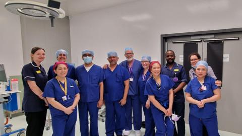 A group of 11 surgical staff in an operating theatre, wearing blue scrubs