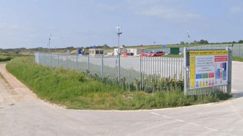 The entrance to a quarry, surrounded by a metal fence, with prefab buildings and parking spaces. To the left, a dirt road and grass bank leads down to the quarry, which can be seen in the distance.