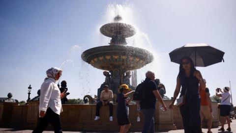 People walk around a fountain, with blue sky and bright sunshine in the background. One woman is holding an umbrella for shade; other people are wearing sunhats and carrying bottles of water.