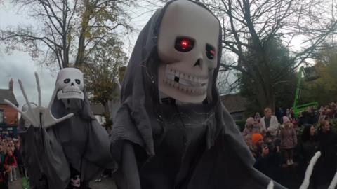 Two lanky skeletons with black capes on are towering above a crowd of people and in various dancing poses. Behind is Lincoln Cathedral in the distance.
